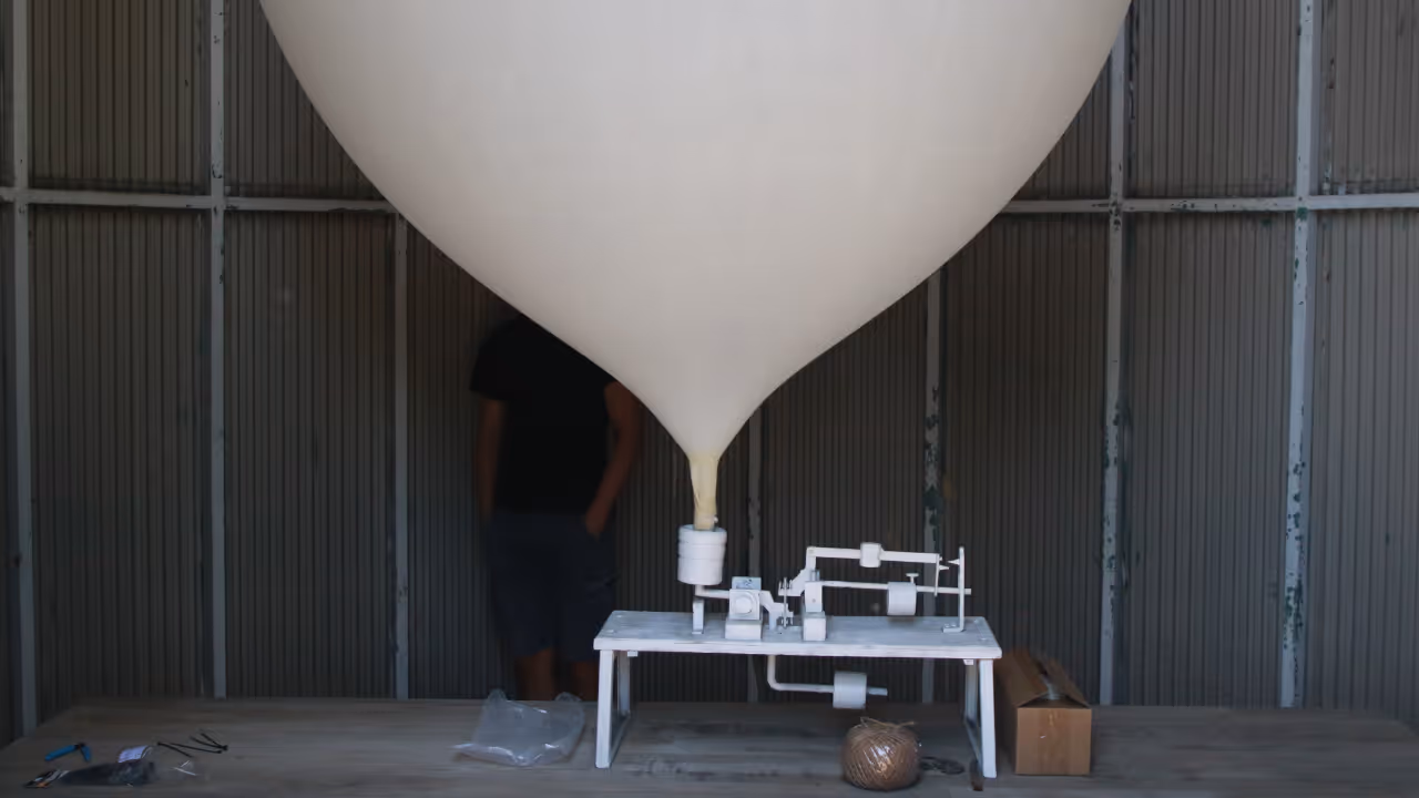 Large weather balloon attached to scientific instruments on a white table inside a metal shed.