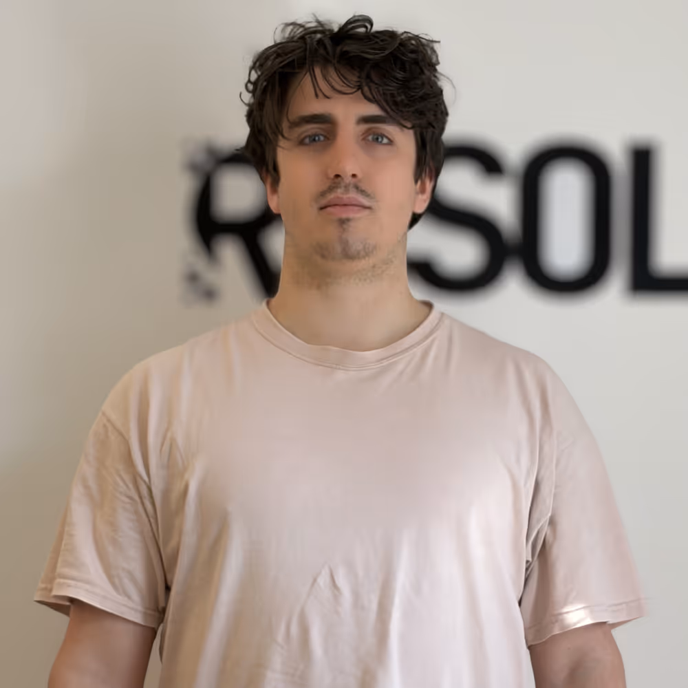 Young man with curly dark hair and blue eyes wearing a light beige t-shirt standing in front of a blurred wall with black text.