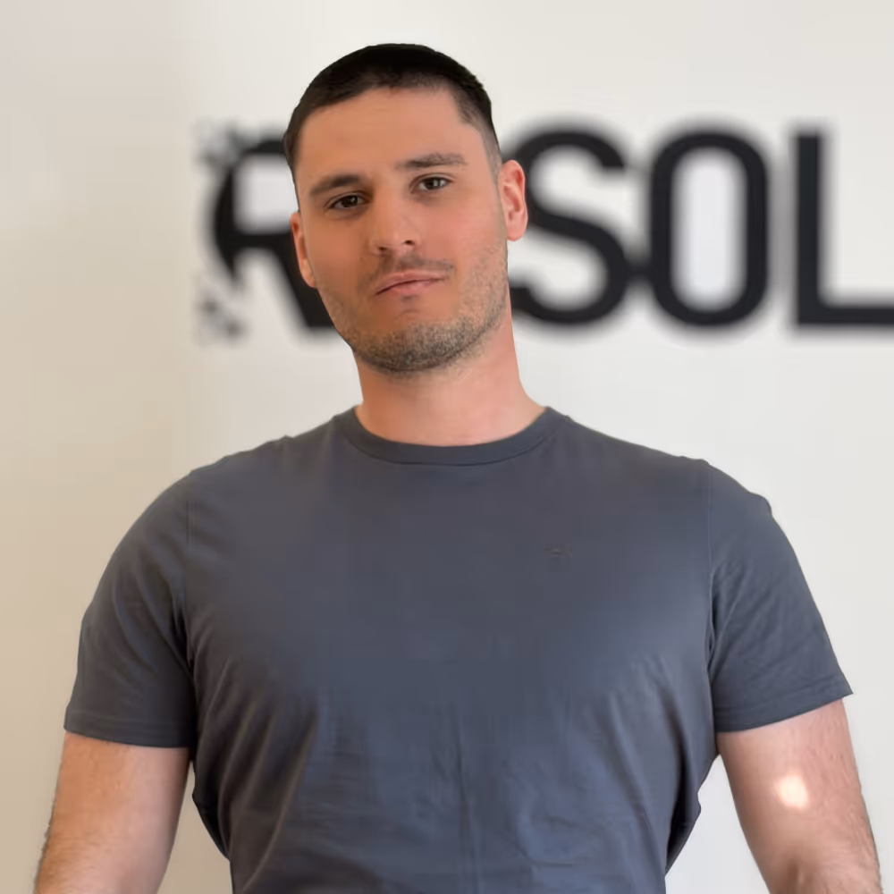 Man with short dark hair wearing a gray t-shirt standing against a light background with blurred text.