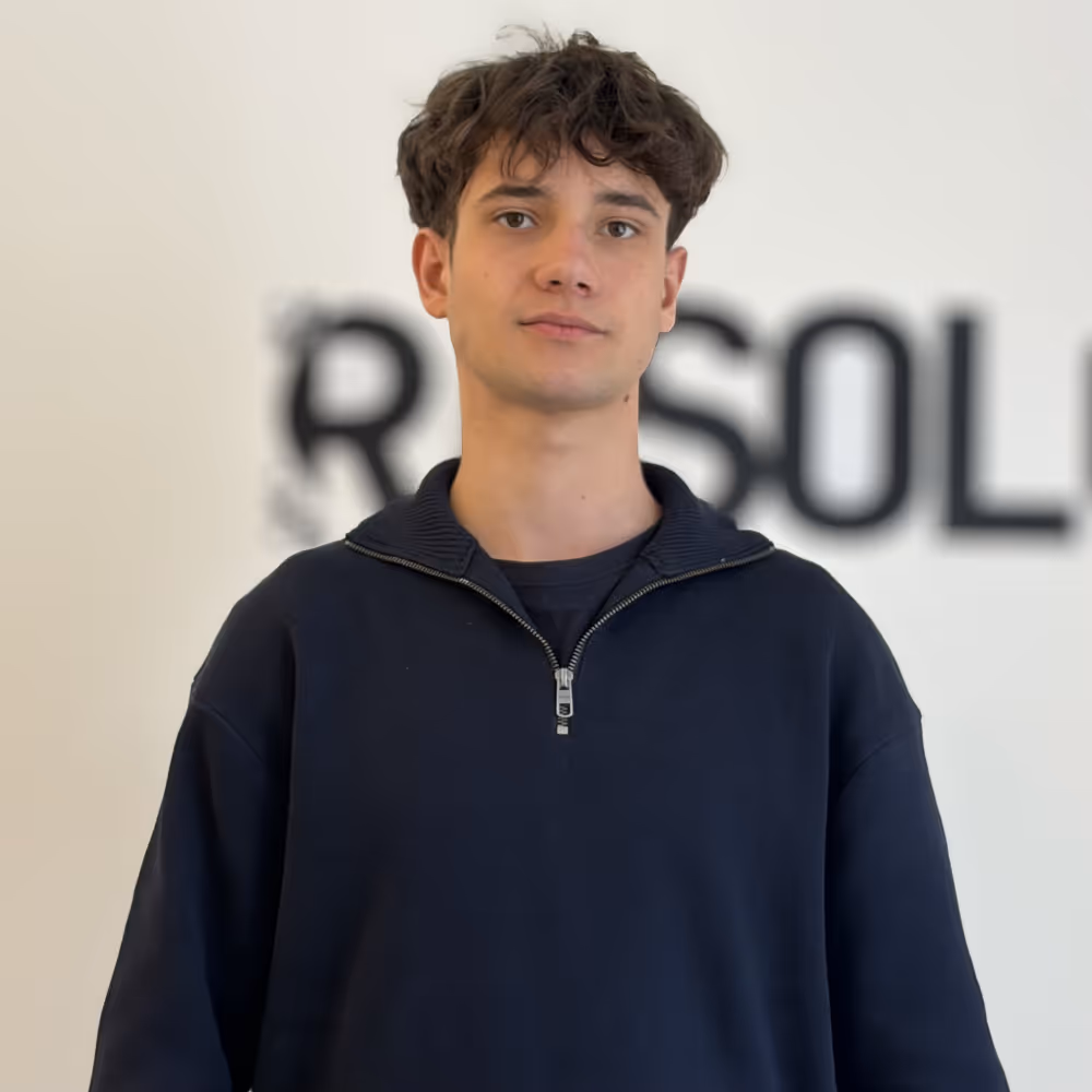 Young man with curly dark hair wearing a navy half-zip sweater standing in front of a blurred white wall with black text.