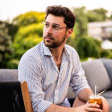 Man with glasses and a striped shirt sitting outdoors holding a drink with a straw.